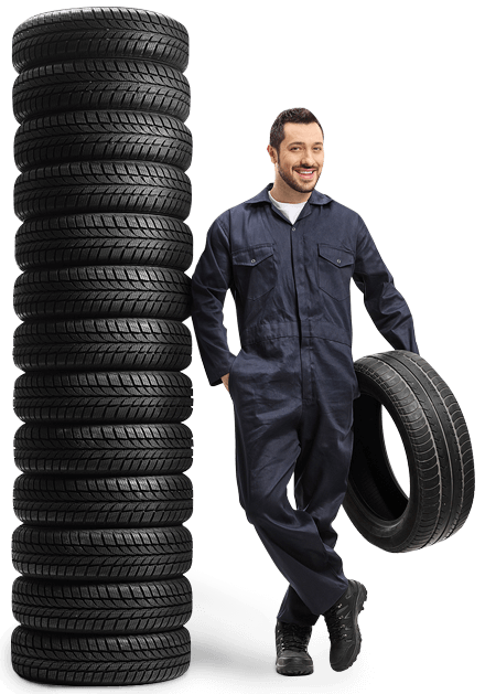 a full shot of an auto mechanic in a dark gray jump suit holding a tire in his left hand, leaning on a stock pile of blacck tires.